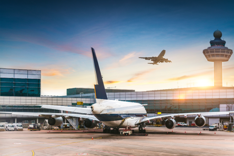 Passenger jets parked at an airport terminal at sunset with another aircraft taking off